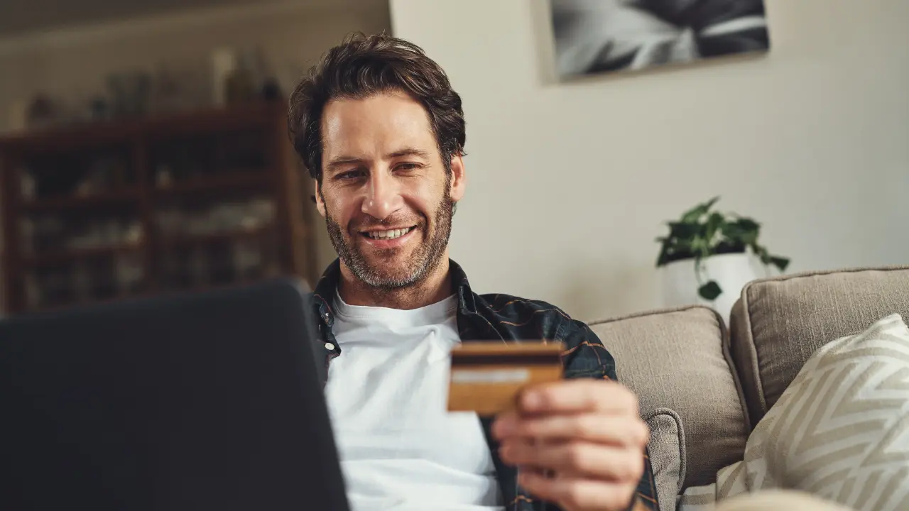 A man uses a laptop and holds a credit card on a couch