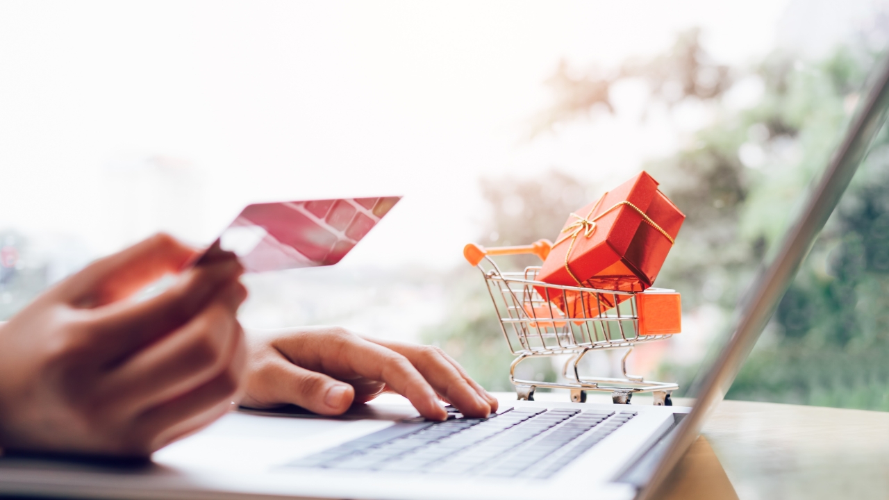 A person enters card details on a laptop beside a small cart with a gift box