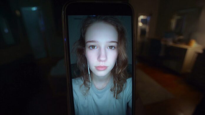 A smartphone displaying a young woman on a video call in a dimly lit room, illustrating the challenge of distinguishing a real caller from a sophisticated deepfake