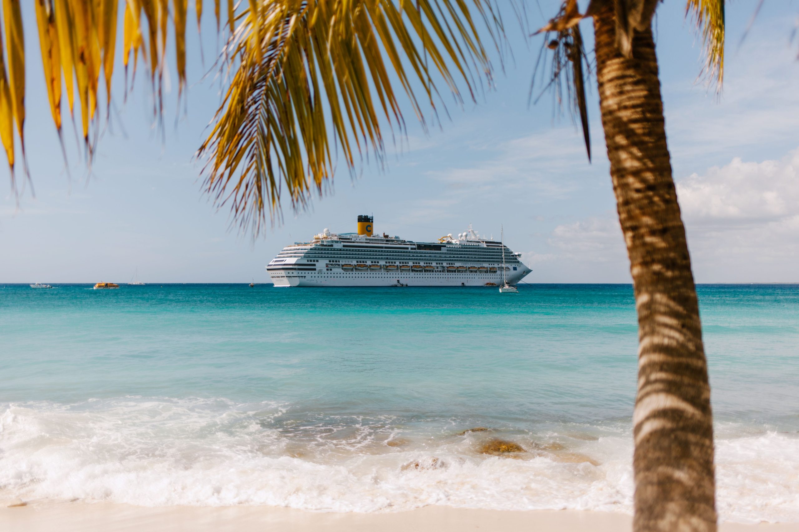 7 Best Cruise Destinations Around the World for 2026 Tropical Caribbean beach with a cruise ship in turquoise water and palm trees.